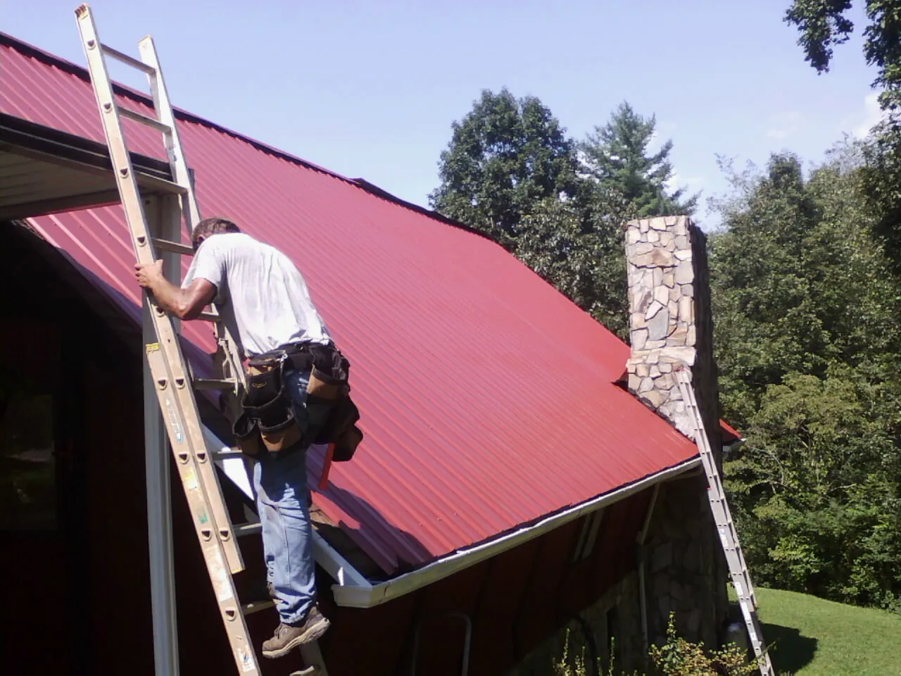 Roofer on a ladder beside a red metal roof and stone chimney — Soffit & Fascia Repair by Roofing Brothers in Lincolnton
